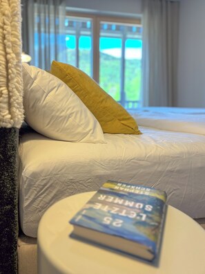 Ein Bett mit weißer Bettwäsche und einem gelben Kopfkissen, ein Buch auf einem Nachttisch und Blick ins Grüne durch ein Fenster.
