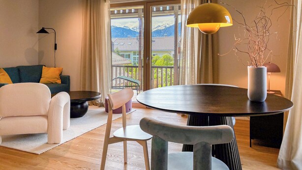 Una sala de estar moderna con una mesa redonda, un sofá, una silla, una lámpara de pie y vistas a las montañas a través de amplios ventanales.