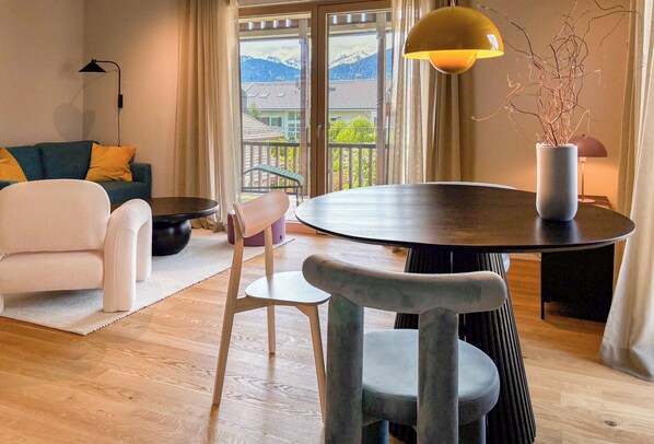 Ein modernes Wohnzimmer mit rundem Tisch, einer Couch, einem Sessel, einer Stehlampe und Blick auf Berge durch große Fenster.