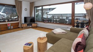 A modern living room with a large window, a flat-screen TV, a sofa, two wooden side tables, and a bookshelf. - Enchanted forest by Loewenherz (Mittenwald)
