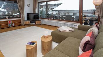Ein modernes Wohnzimmer mit großem Fenster, einem Flachbildfernseher, einer Couch, zwei Holztischen und einem Bücherregal.