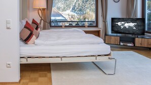 A bedroom with a bed, a TV, a window with a view of snow-covered mountains, and a lamp. - Enchanted forest by Loewenherz (Mittenwald)