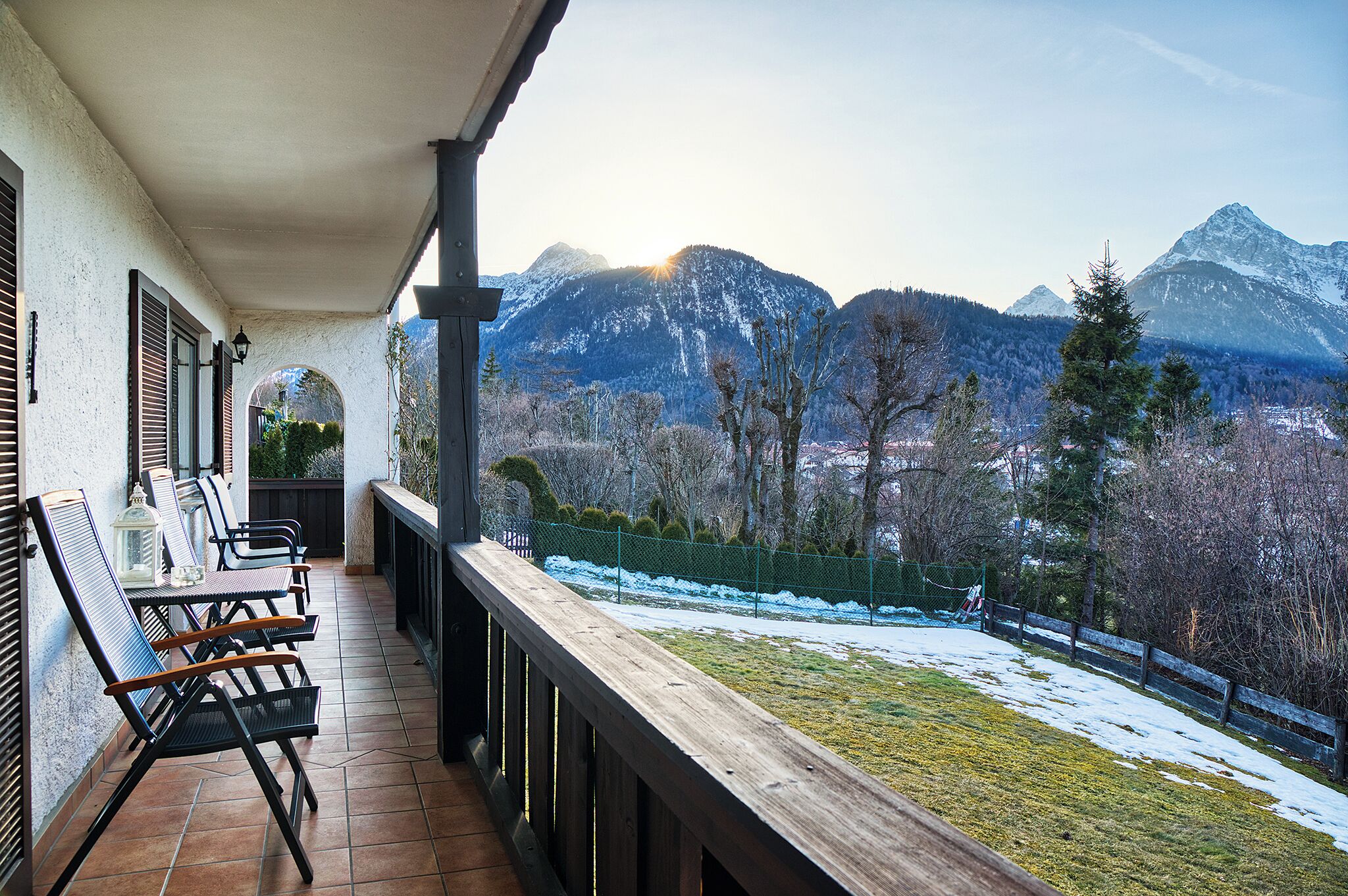 Ein Balkon mit Gartenmöbeln, Blick auf verschneite Berge und ein eingezäunter Garten.
