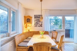 Ein Essbereich mit Holztisch und Bänken, einer Wandleuchte und Blick auf Bäume durch die Fenster.