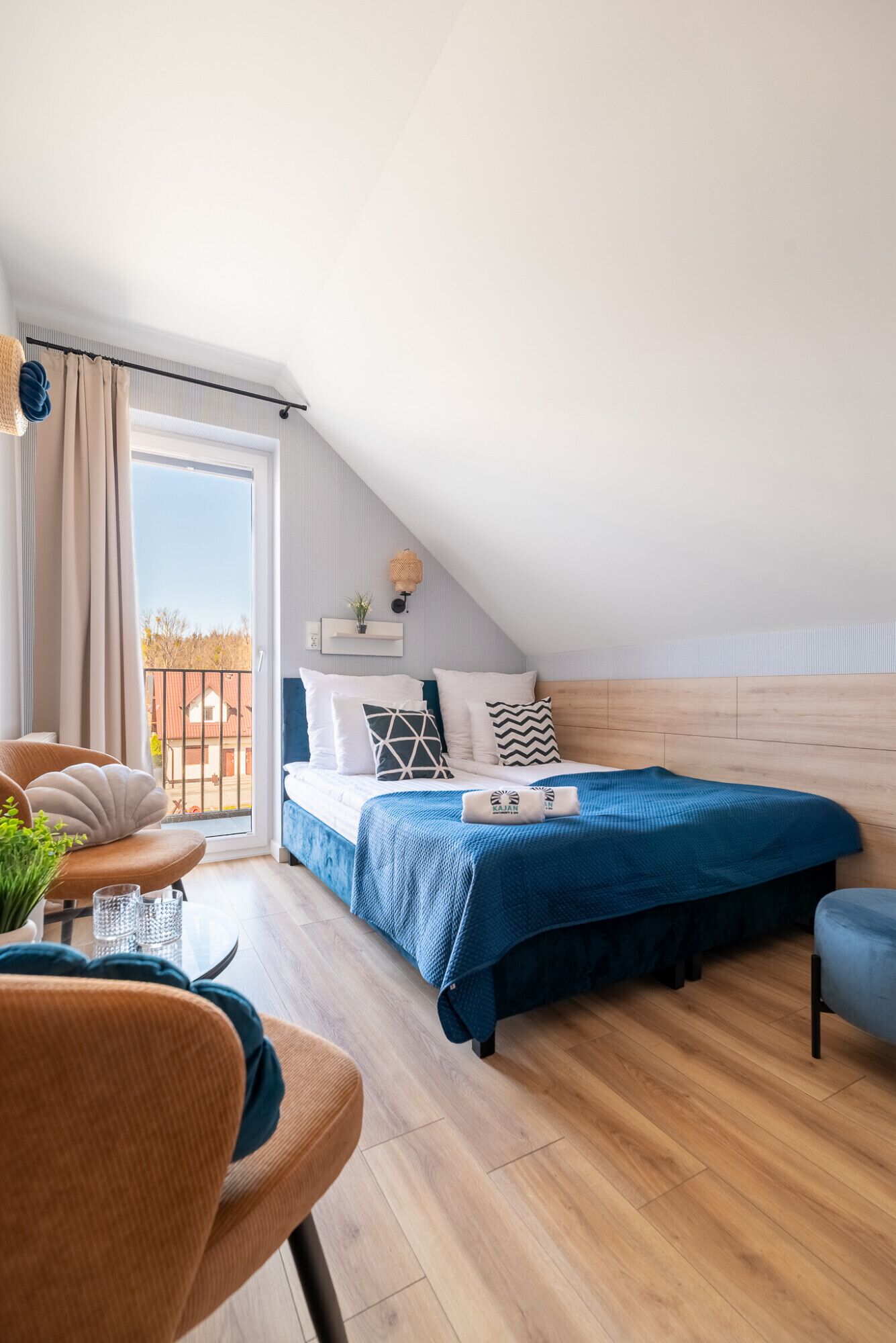 Quarto moderno com teto inclinado, uma cama grande com colcha azul, cabeceira de madeira e varanda com vista.