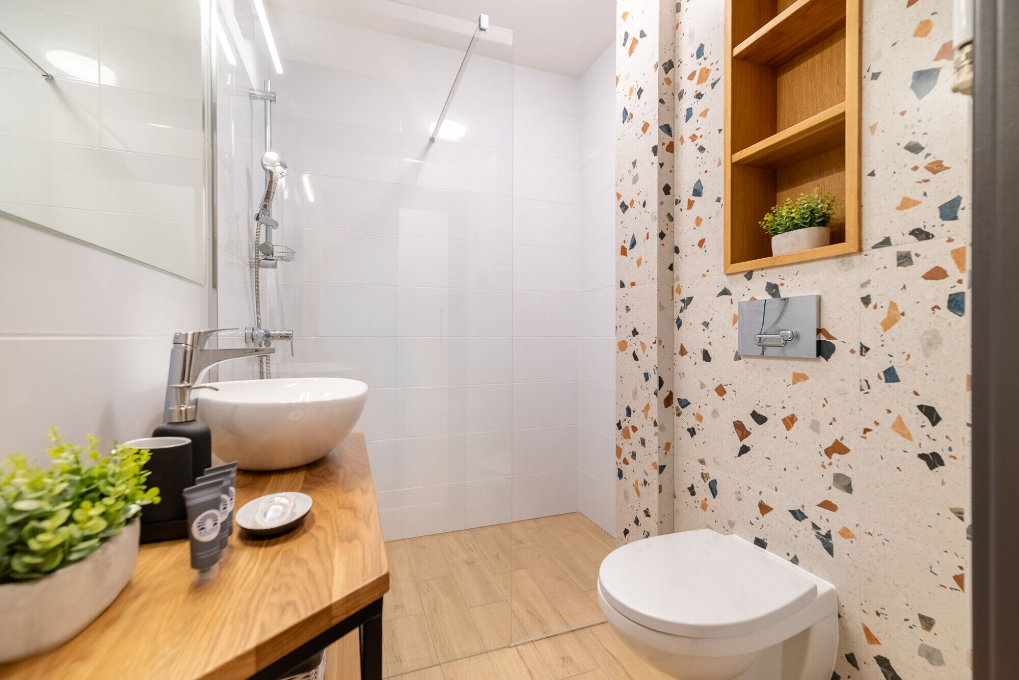 Een badkamer met een houten wastafelmeubel, twee wastafels, een toilet, een douche en een wand met terrazzo patroon.