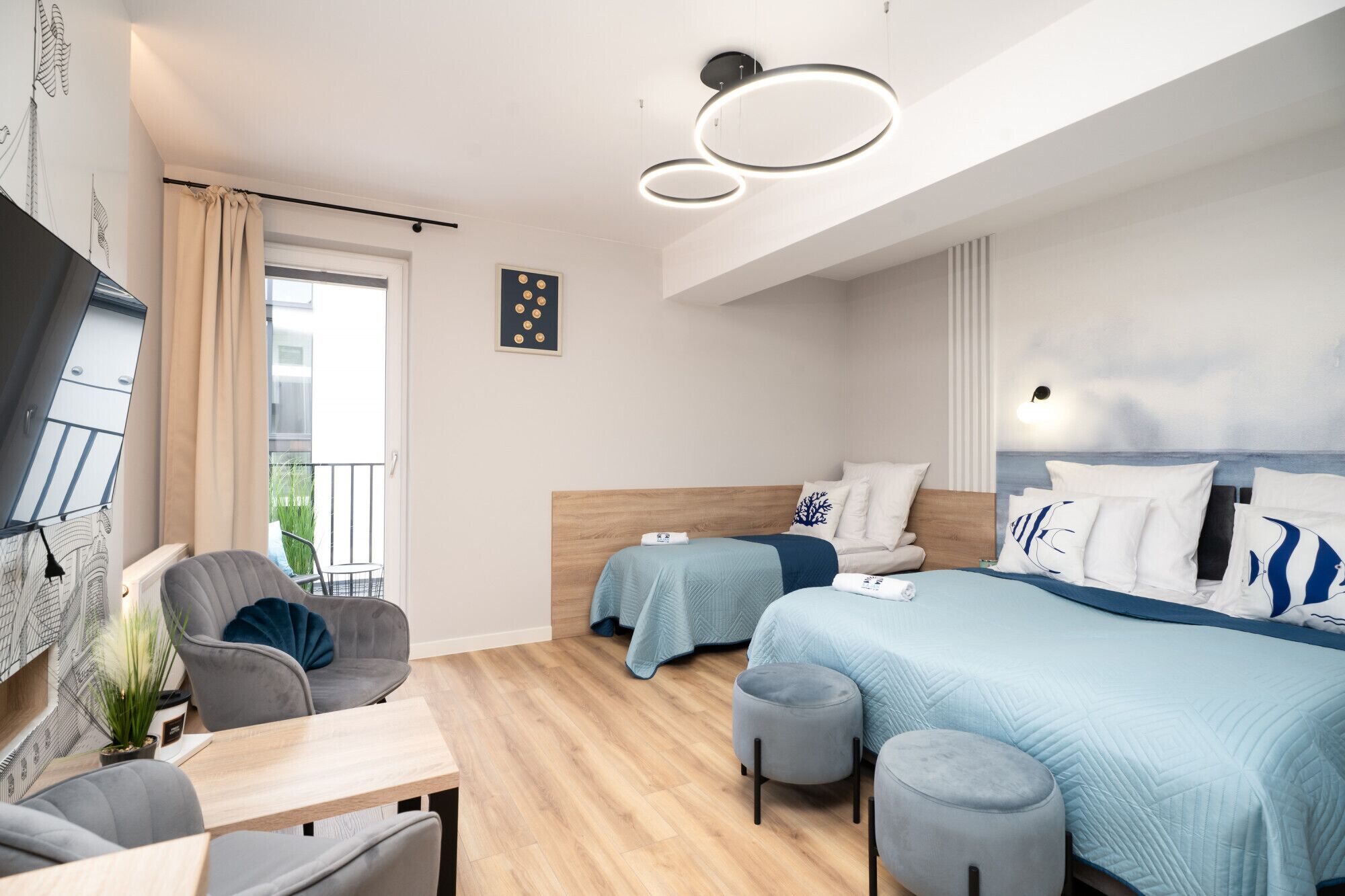 A modern hotel room with two beds, a sitting area with a table and chairs, a balcony with curtains, and a ceiling light fixture.