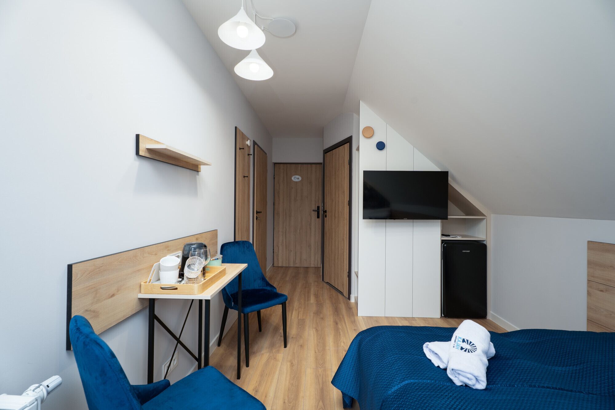 A compact hotel room with a bed, a desk, a chair, a small kitchenette, and a wardrobe.