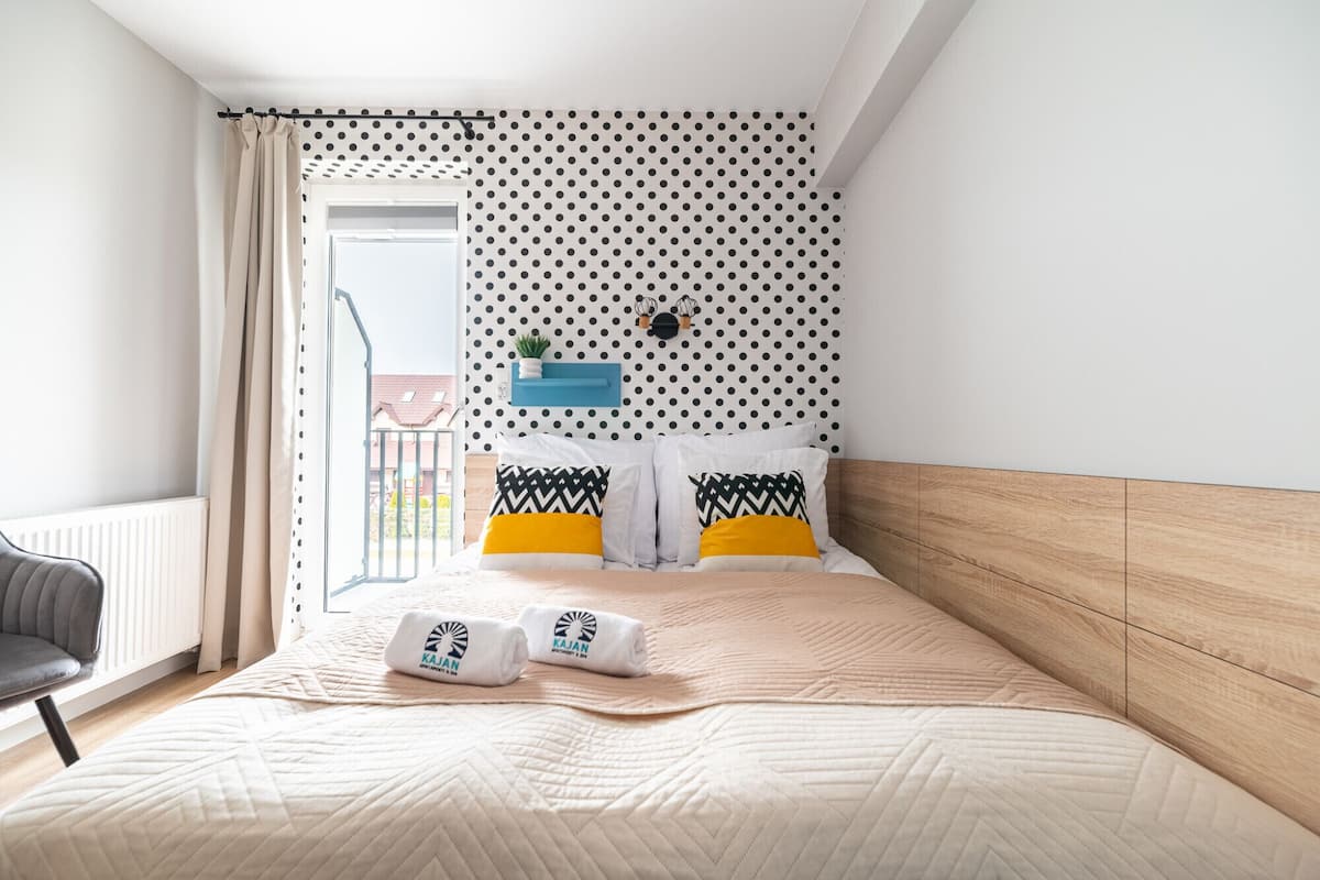 Una habitación de hotel con cama, una silla, una ventana con cortinas, un papel tapiz con lunares y un estante pequeño con una planta.