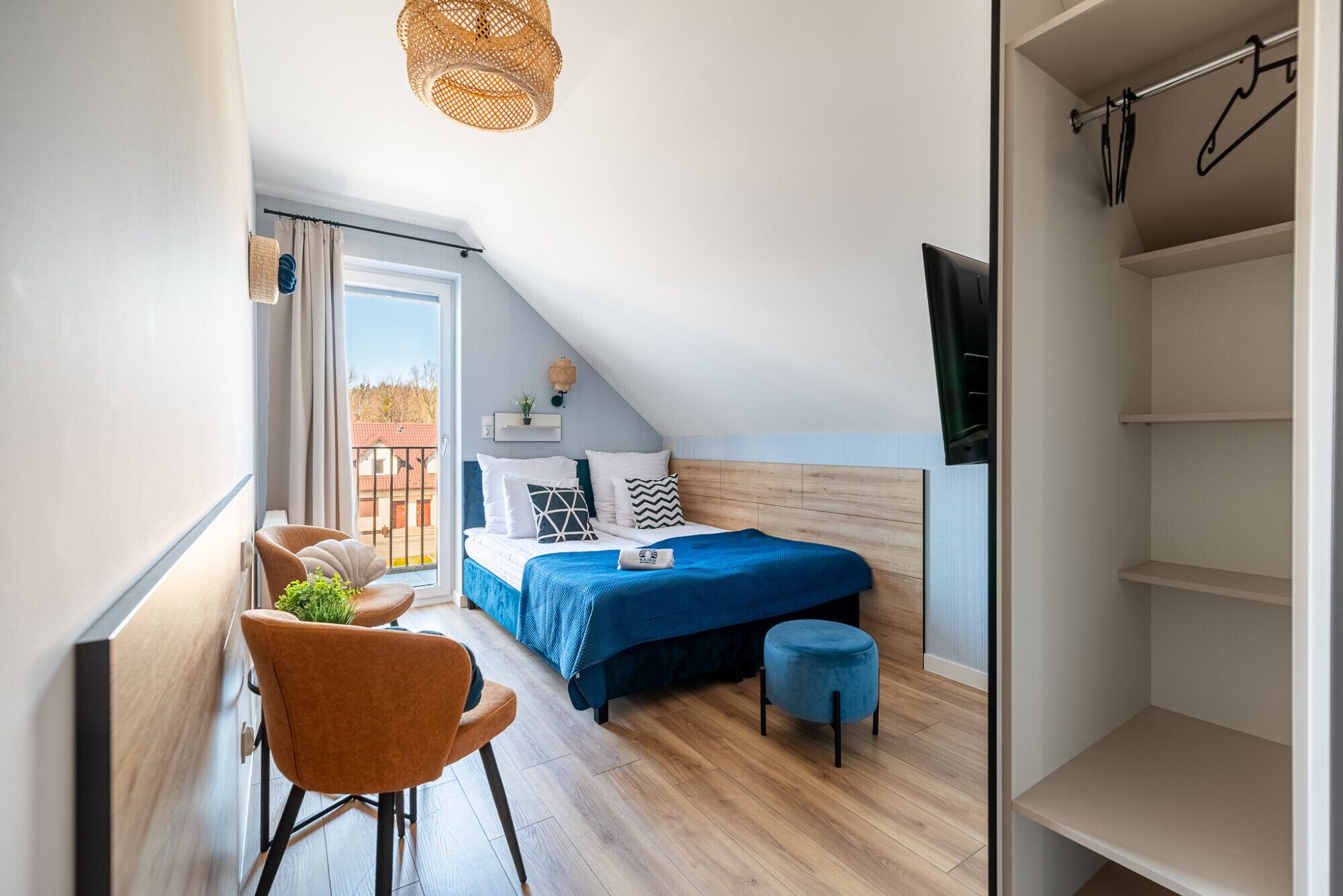 A bedroom with a bed, a chair, a small stool, a hanging light fixture, and a window with a view.