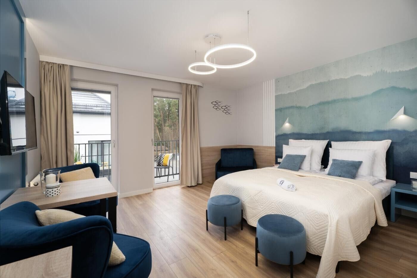 Habitación de hotel con cama, escritorio, dos otomanas, una silla, televisor y balcón con cortinas.