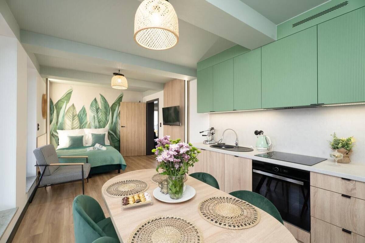 Una cocina moderna con armarios verdes, un comedor con una mesa para dos, y un dormitorio con un gran mural de plantas.
