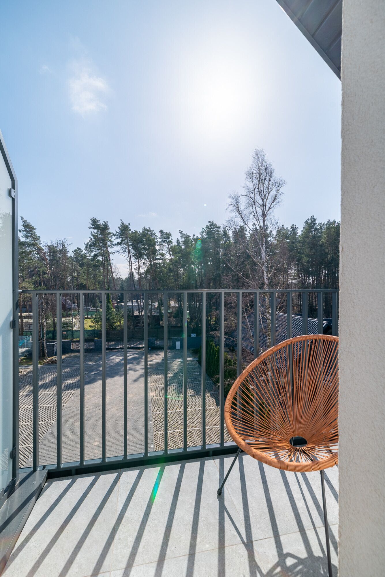 A balcony with a metal railing, a wicker chair, and a view of a parking area and trees.
