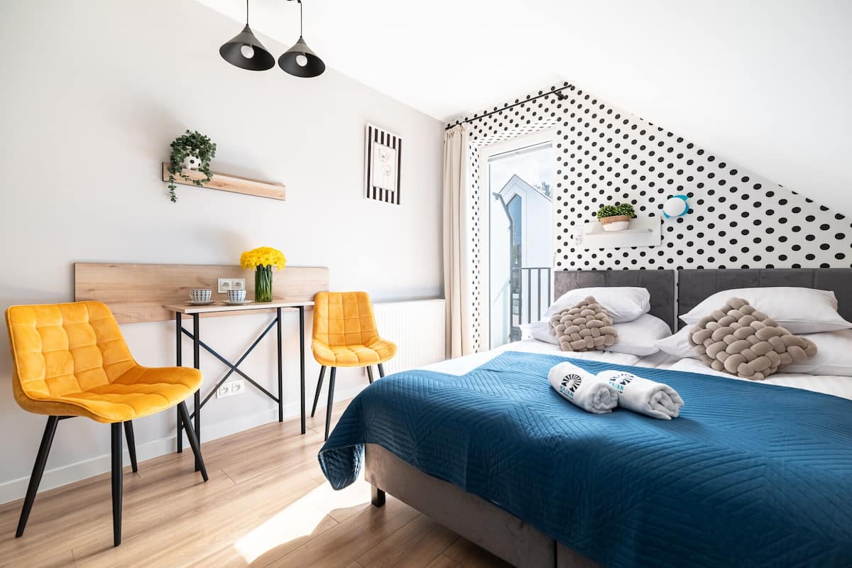 Un dormitorio moderno con una cama, dos sillones, una mesita, una estantería con una planta y un ventanal con vistas.