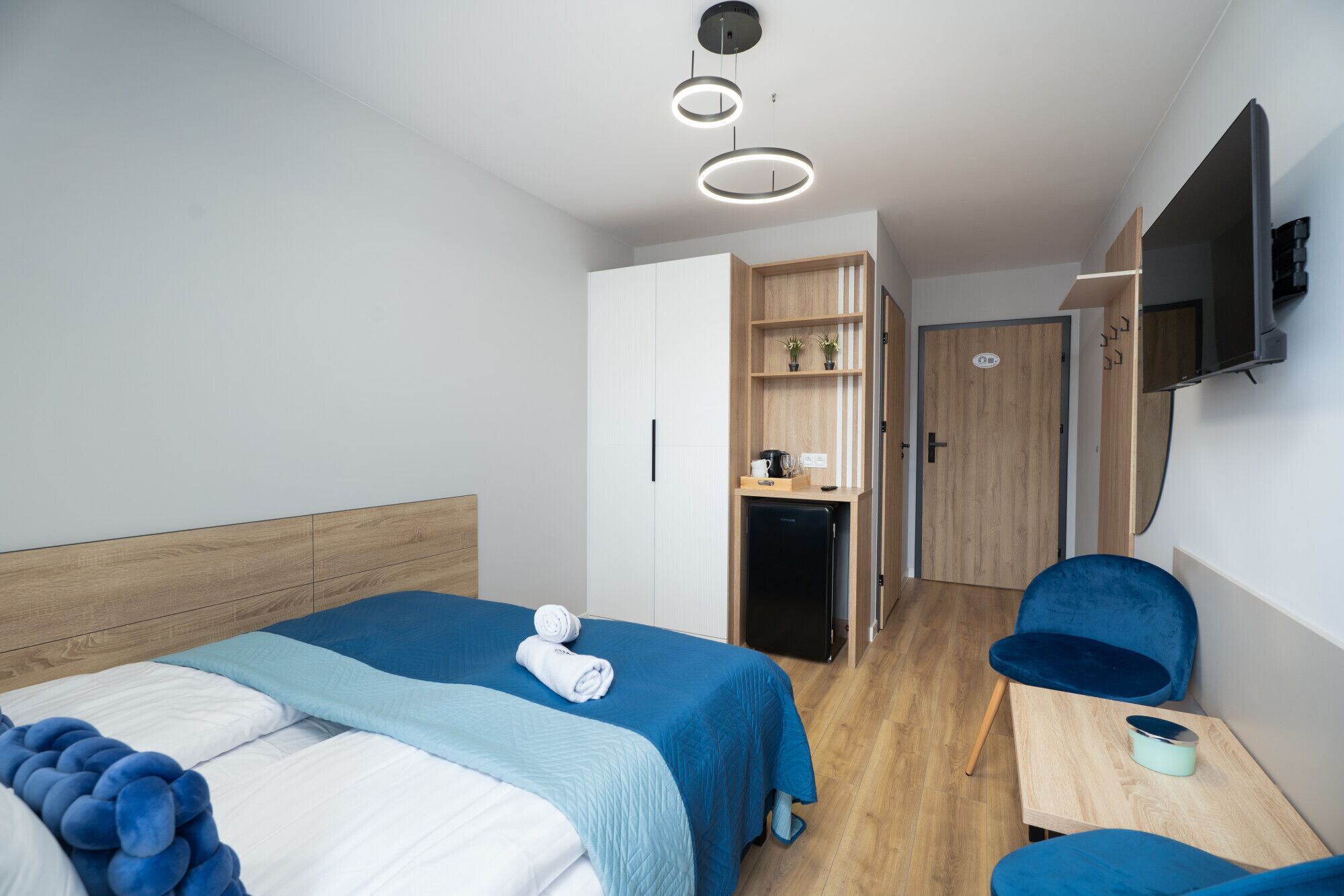 A hotel room with two beds, a desk, a chair, a TV, and a kitchenette.