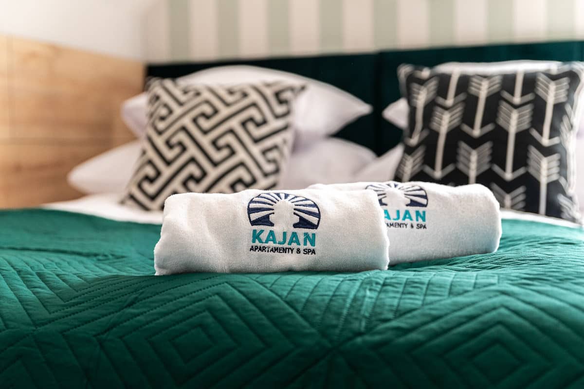 Una cama con un edredón verde, dos toallas enrolladas con el logotipo 'Kajan' y almohadas estampadas.