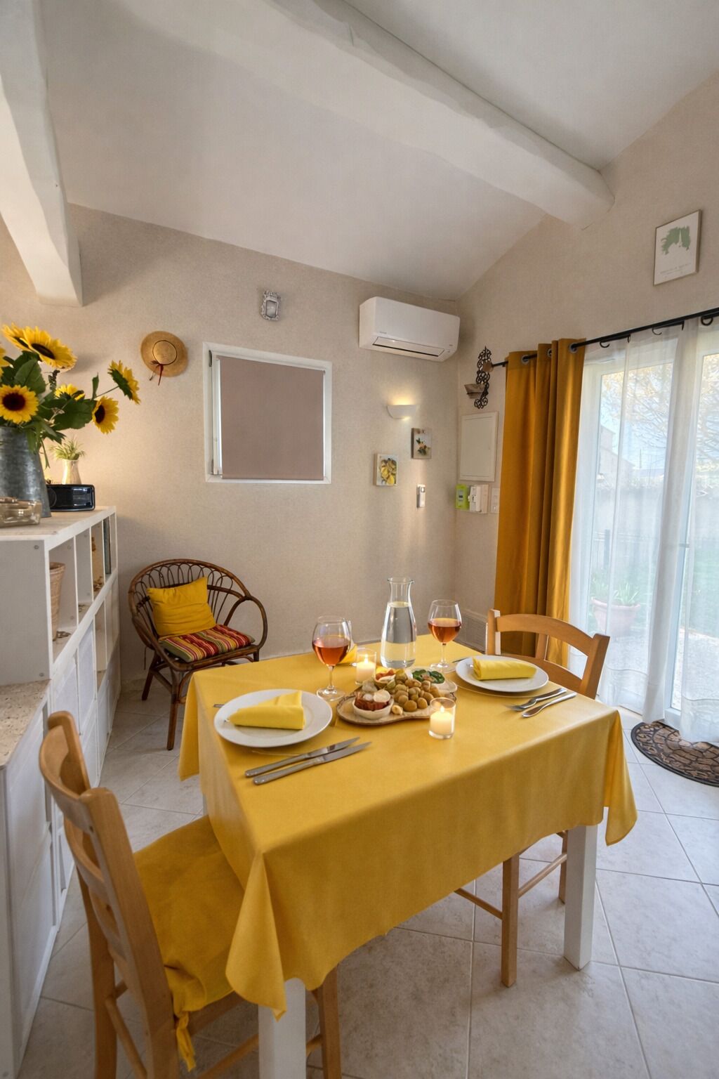 Een eetgedeelte met een tafel gedekt voor twee personen, een rieten stoel, een zonnehoed aan de muur en een raam met gordijnen.