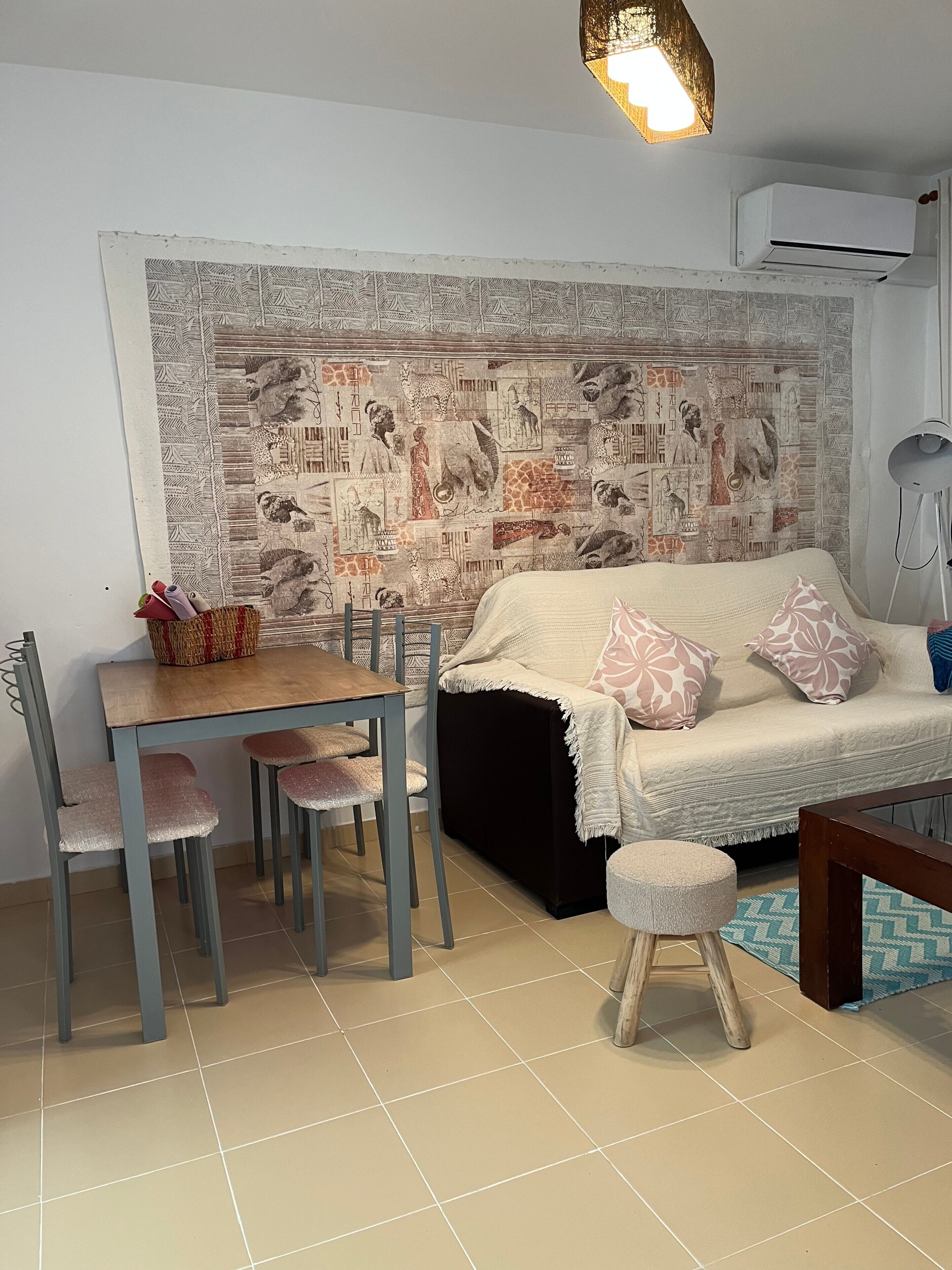 A room with a dining table, chairs, a sofa, a small stool, and a wall tapestry.