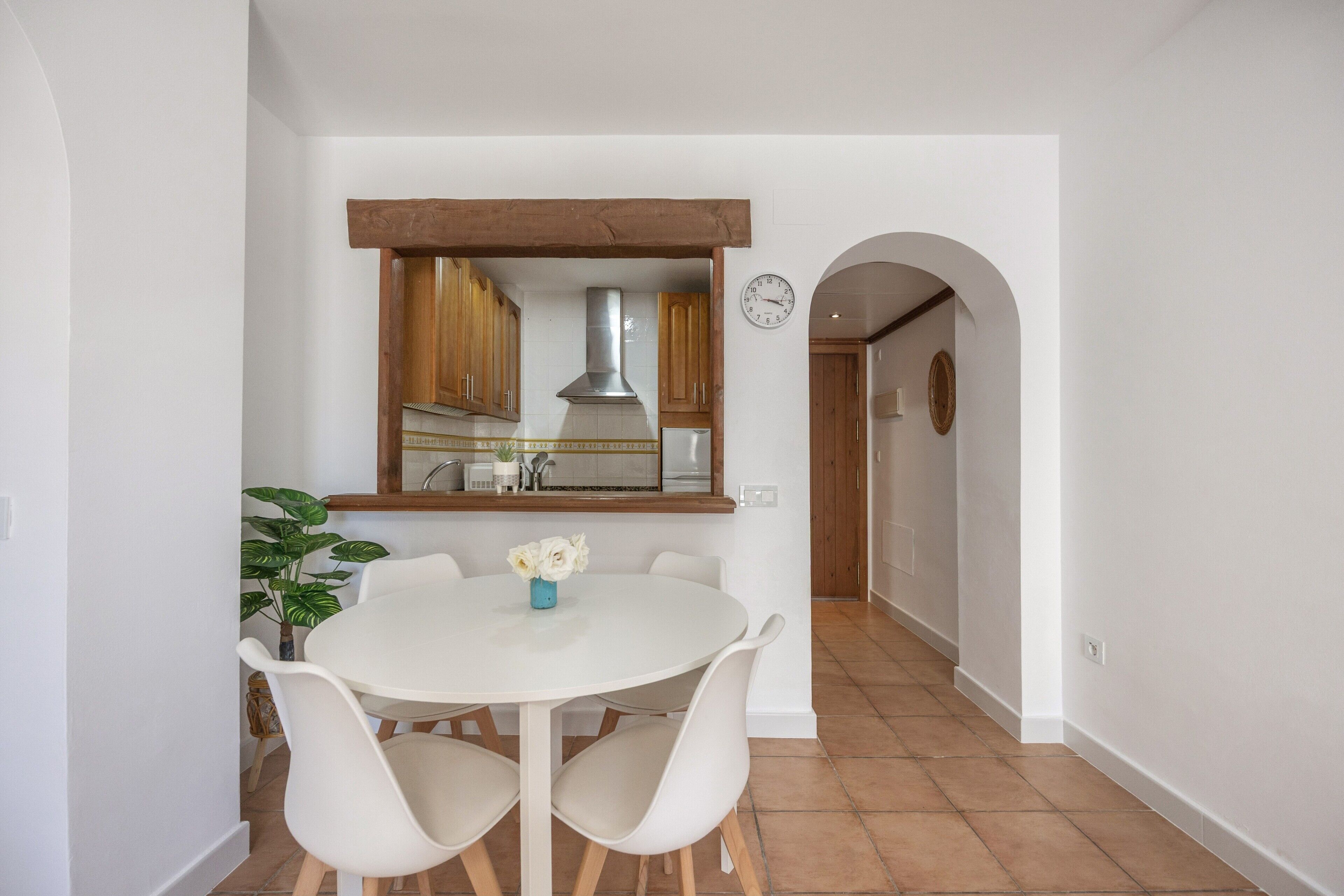 Een eetruimte met een witte tafel en stoelen, een kamerplant en een keuken op de achtergrond.