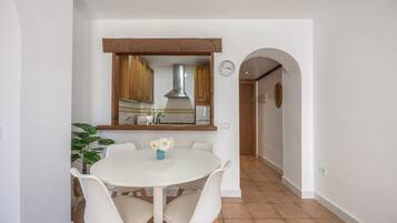 Een eetruimte met een witte tafel en stoelen, een kamerplant en een keuken op de achtergrond.
