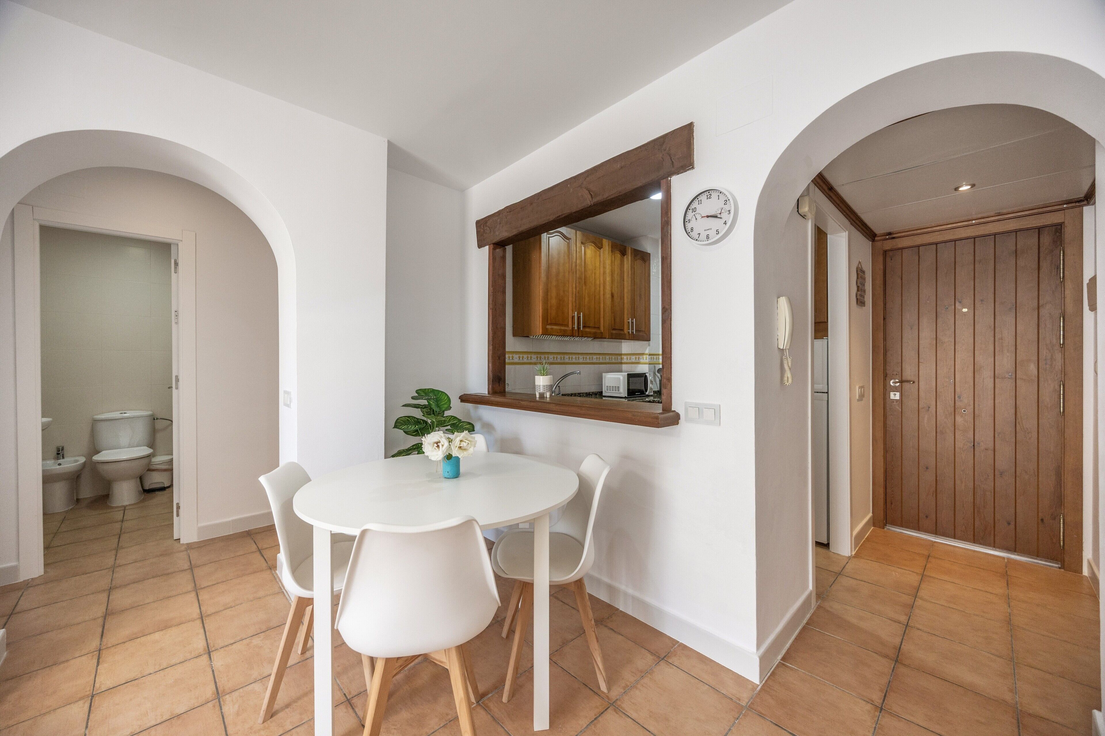 Een eetgedeelte met een ronde tafel en stoelen, een spiegel met houten lijst en een klok aan de muur.