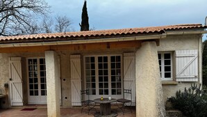 Een landelijk gebouw met een pannendak, grote ramen en een overdekt terras met een tafel en stoelen.