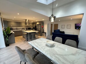 A modern kitchen with a central island, bar stools, a dining table with chairs, a sofa, and a TV mounted on the wall.