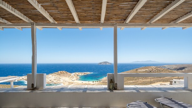 Un balcón con vistas al mar y a las montañas, una pared blanca y dos tumbonas.