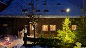 Un espacio exterior cubierto de nieve, con una valla de madera, un árbol de Navidad con luces y un edificio con ventanas.