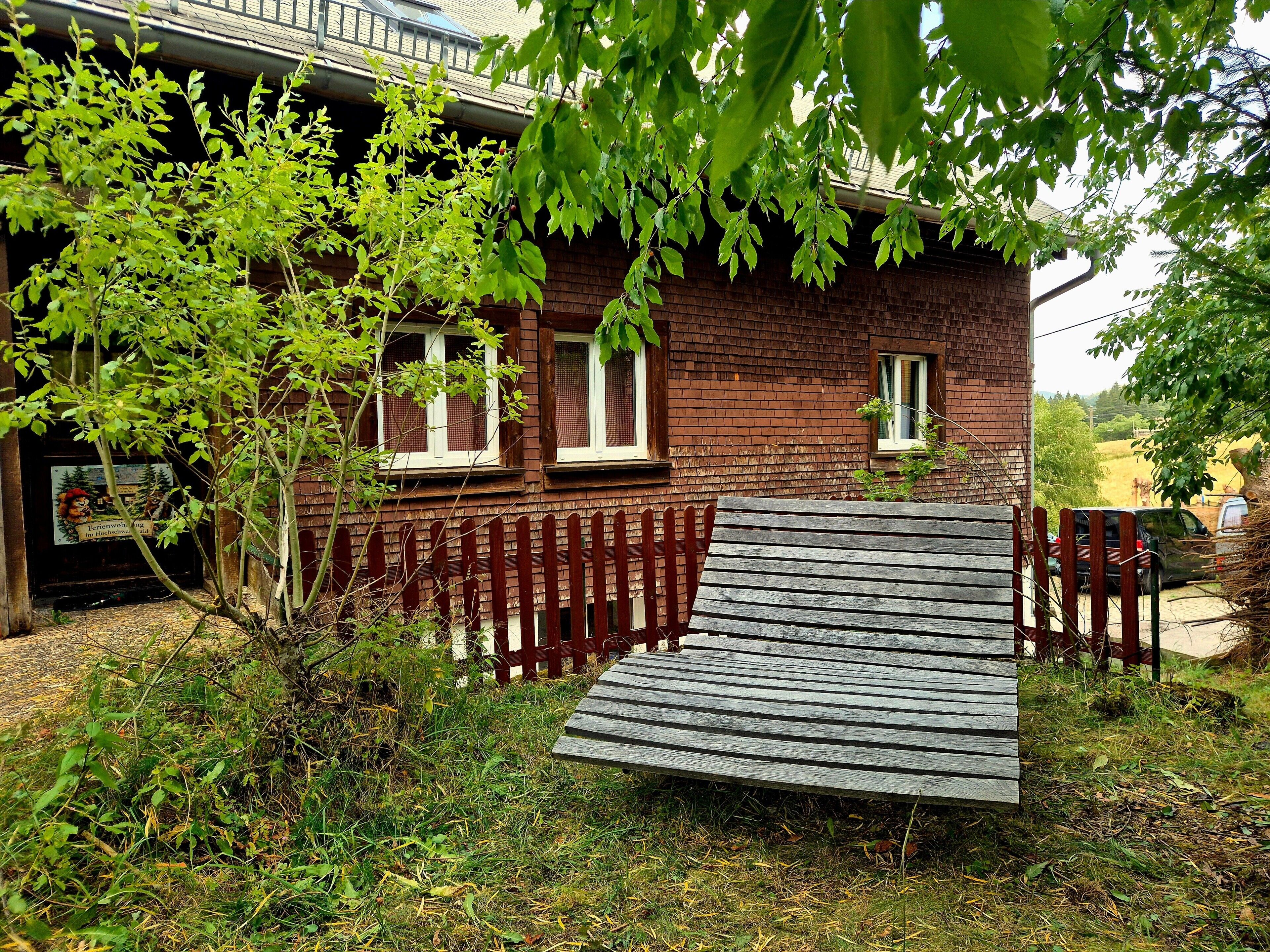 A wooden bench in front of a brick house with a red fence and a tree with green leaves.