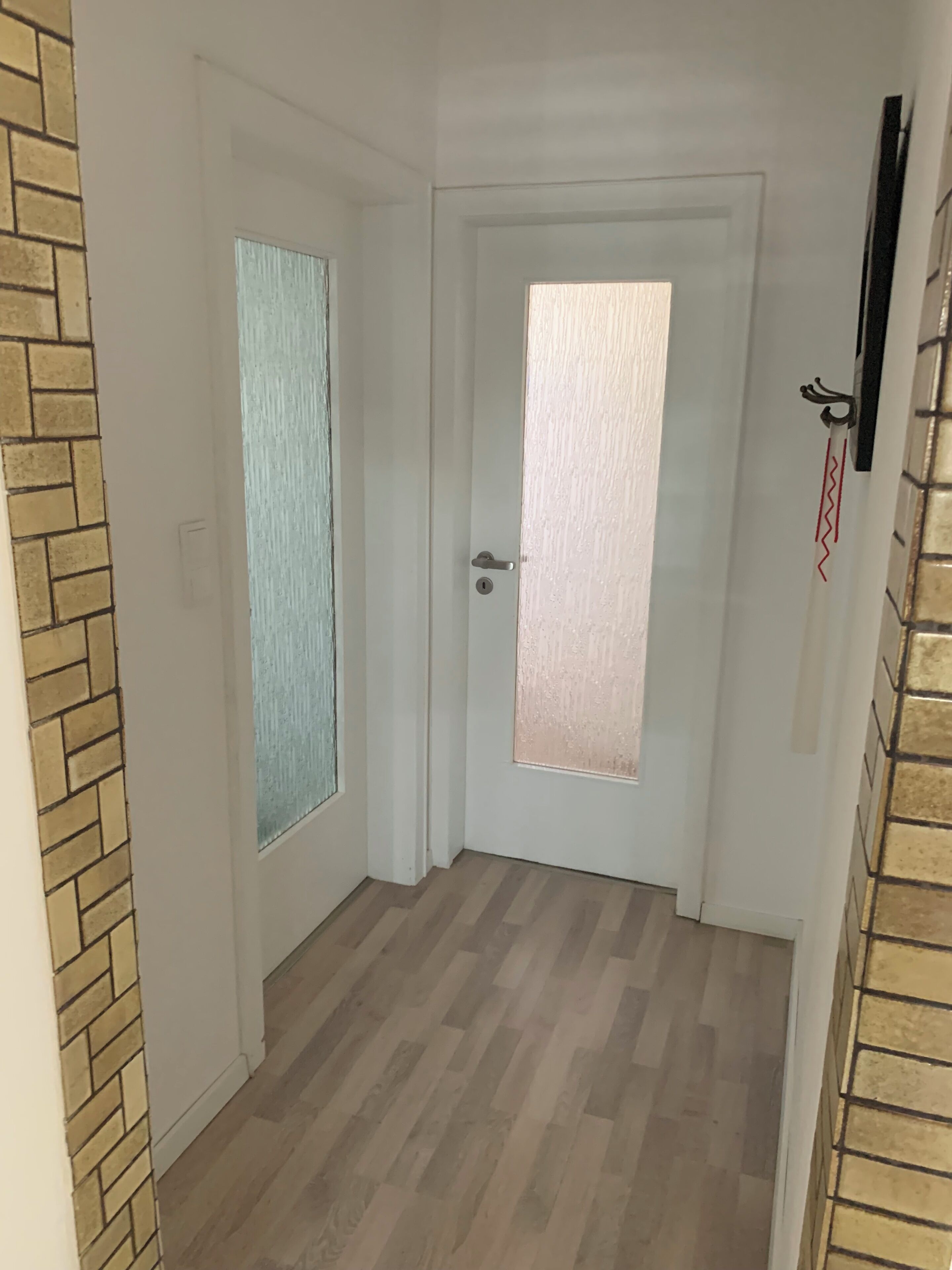 Un couloir avec deux portes : l’une est dotée d’une vitre dépolie et d’un trou de serrure, et l’autre, d’une poignée et d’un trou de serrure. Le sol est en bois et un mur de briques se trouve sur l’un des côtés.