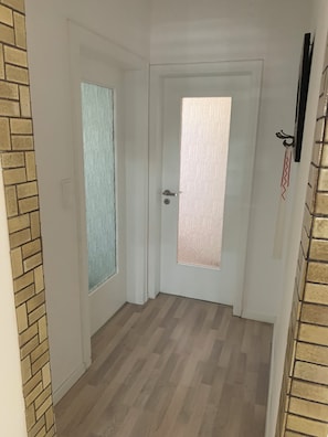 Ein Flur mit zwei Türen, eine mit satiniertem Glas und Schlüsselloch, die andere mit Griff und Schlüsselloch. Der Boden ist Holzboden, und an einer Seite ist eine Ziegelwand.