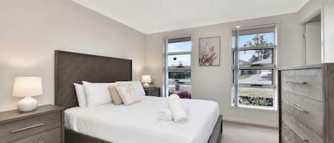 A modern bedroom with a large bed, bedside tables, a dresser, and a window with a view.