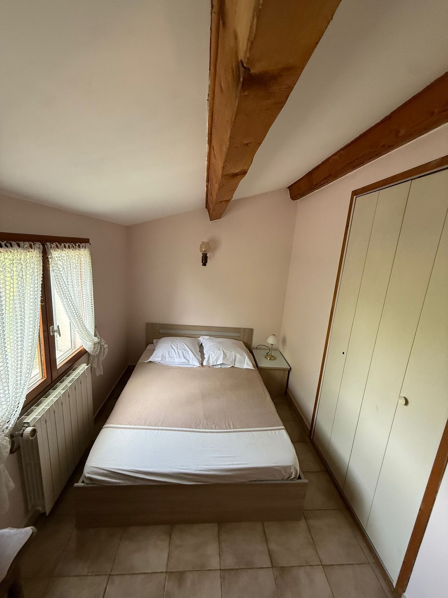 A bedroom with a bed, a nightstand, a radiator, a window with curtains, and a wardrobe.