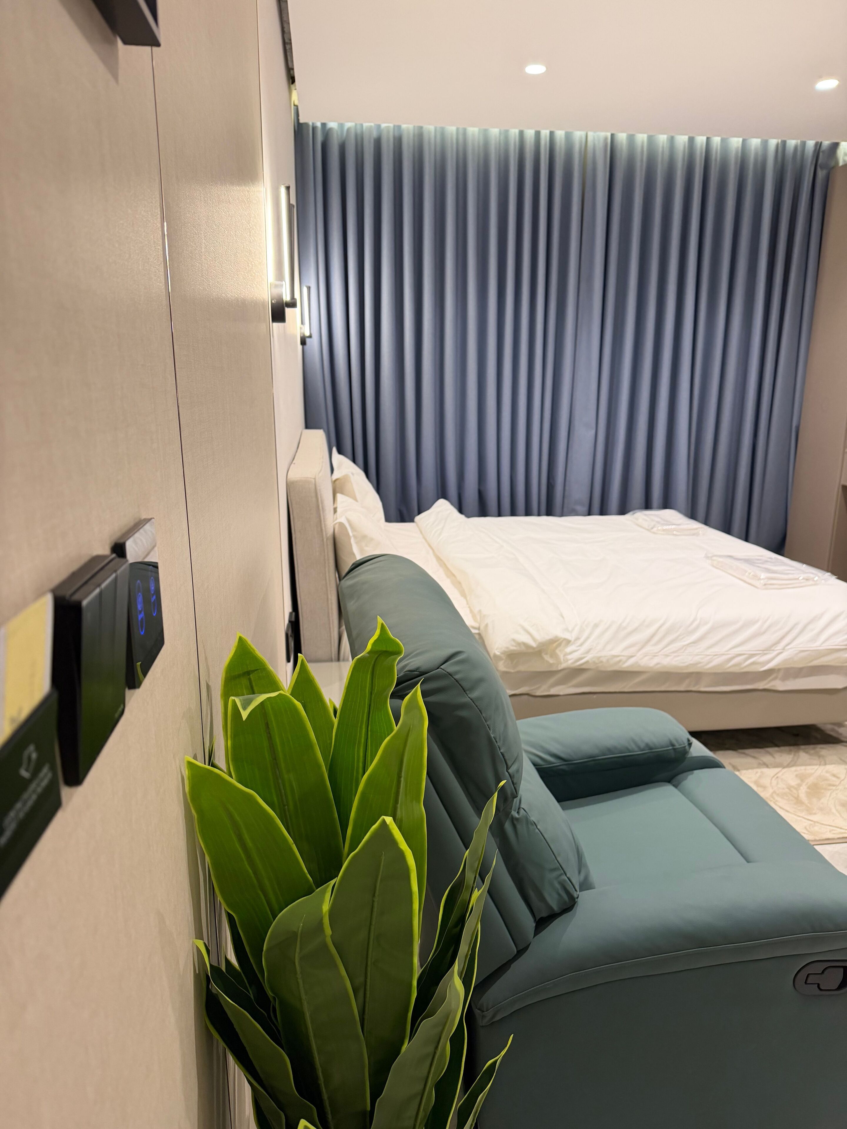 A modern hotel room with a bed, a massage chair, a wall-mounted control panel, and a large window with curtains.