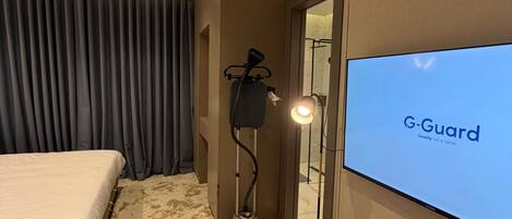 A hotel room with a bed, a flat-screen TV displaying 'G-Guard,' a hairdryer, and an air conditioning unit.