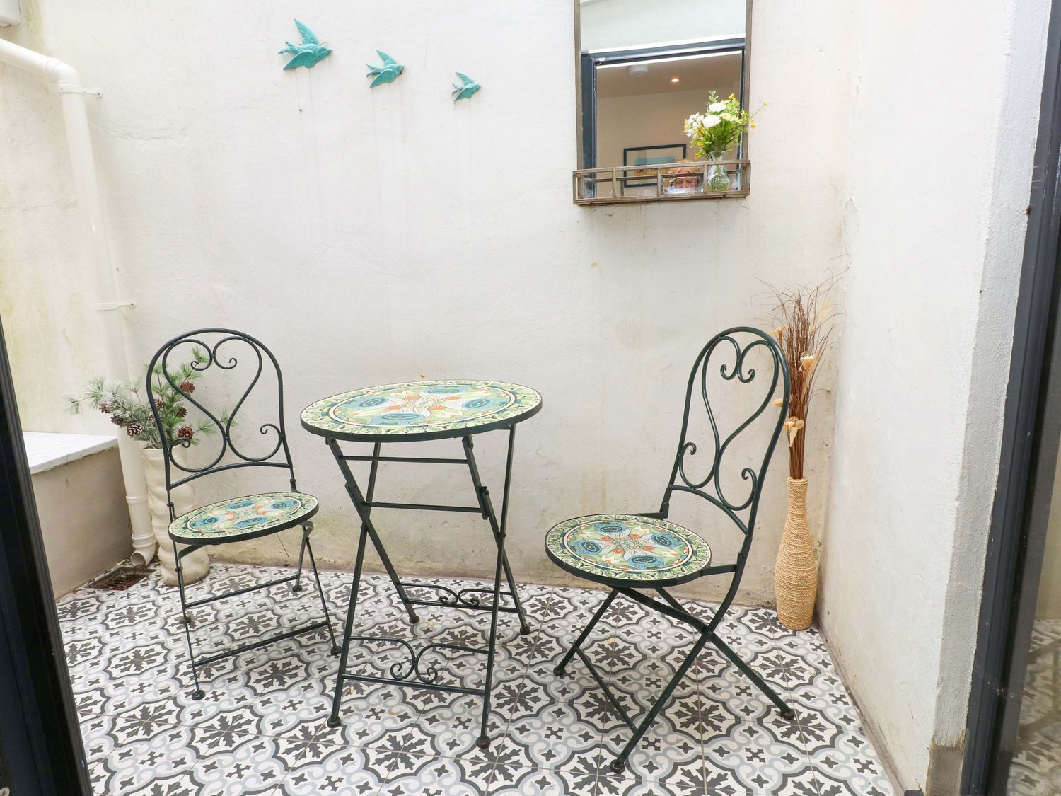 A small patio with two metal chairs and a table, a tiled floor, a white wall, and a window with a mirror and a vase of flowers.