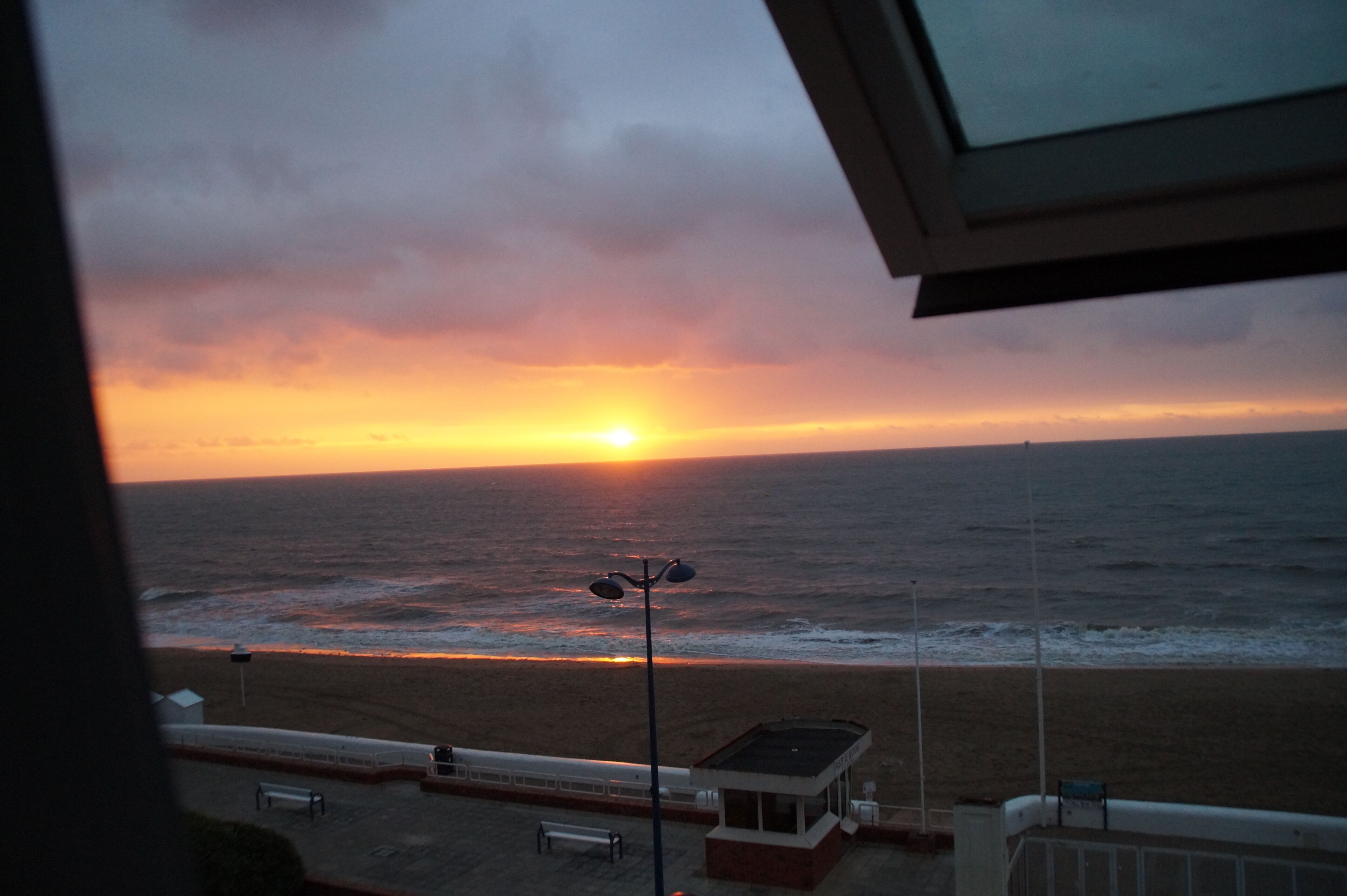 Une vue sur le coucher de soleil, prise depuis une fenêtre donnant sur une plage, avec un banc et un petit bâtiment.