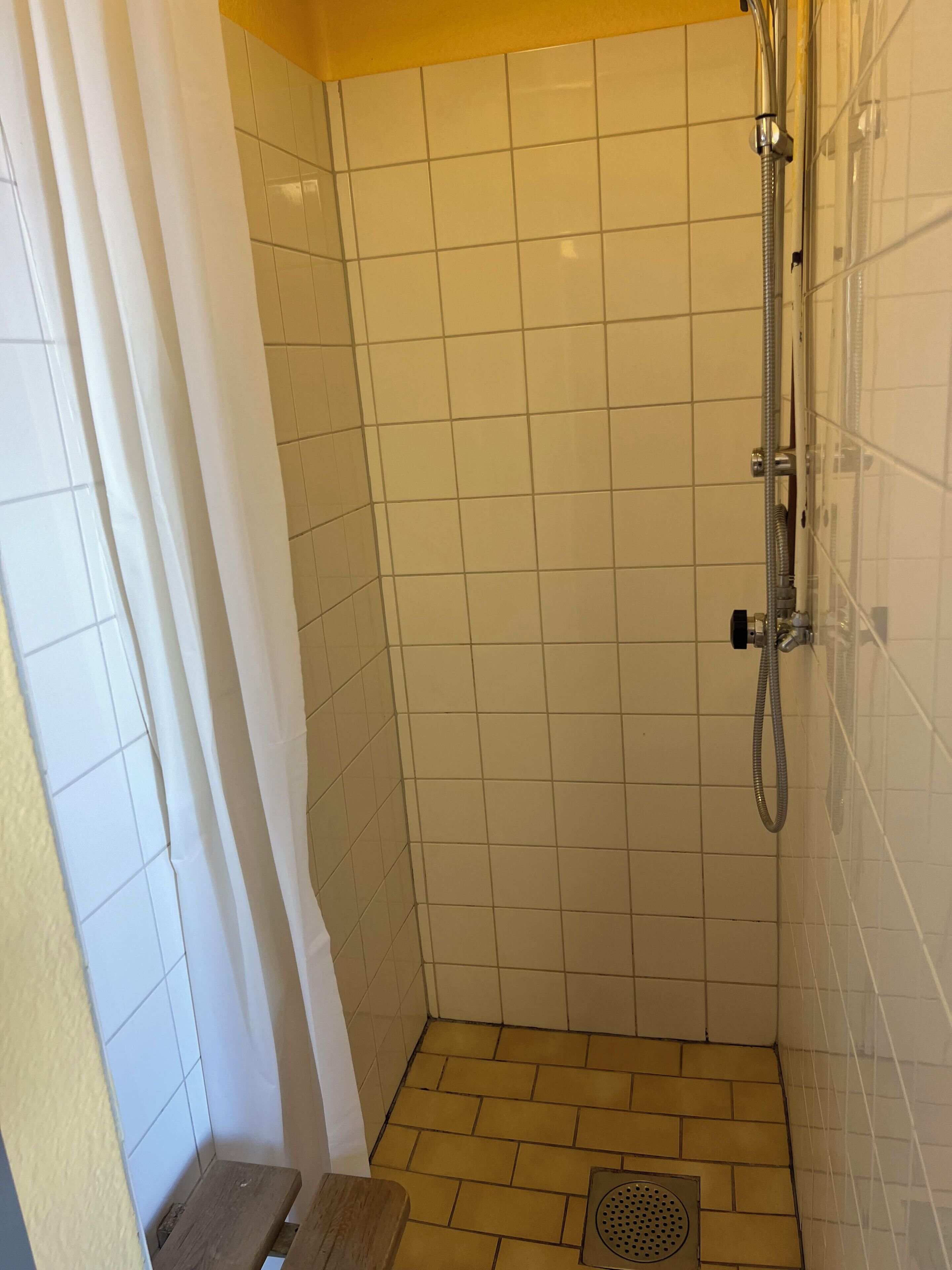 A small, tiled shower room with a showerhead, a drain, and a wooden bench.