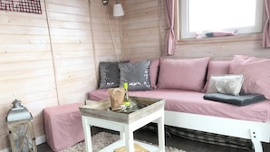 Ein gemütliches Zimmer mit einer Holzwand, einem weißen Sofa mit roten und weißen Karokissen, einem kleinen weißen Tisch mit einer Flasche und einer Tasche sowie einem flauschigen weißen Teppich.