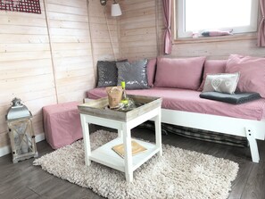 Une pièce chaleureuse avec un mur en bois, un canapé blanc orné de coussins à carreaux rouges et blancs, une petite table blanche sur laquelle se trouvent une bouteille et un sac, et un tapis blanc moelleux.