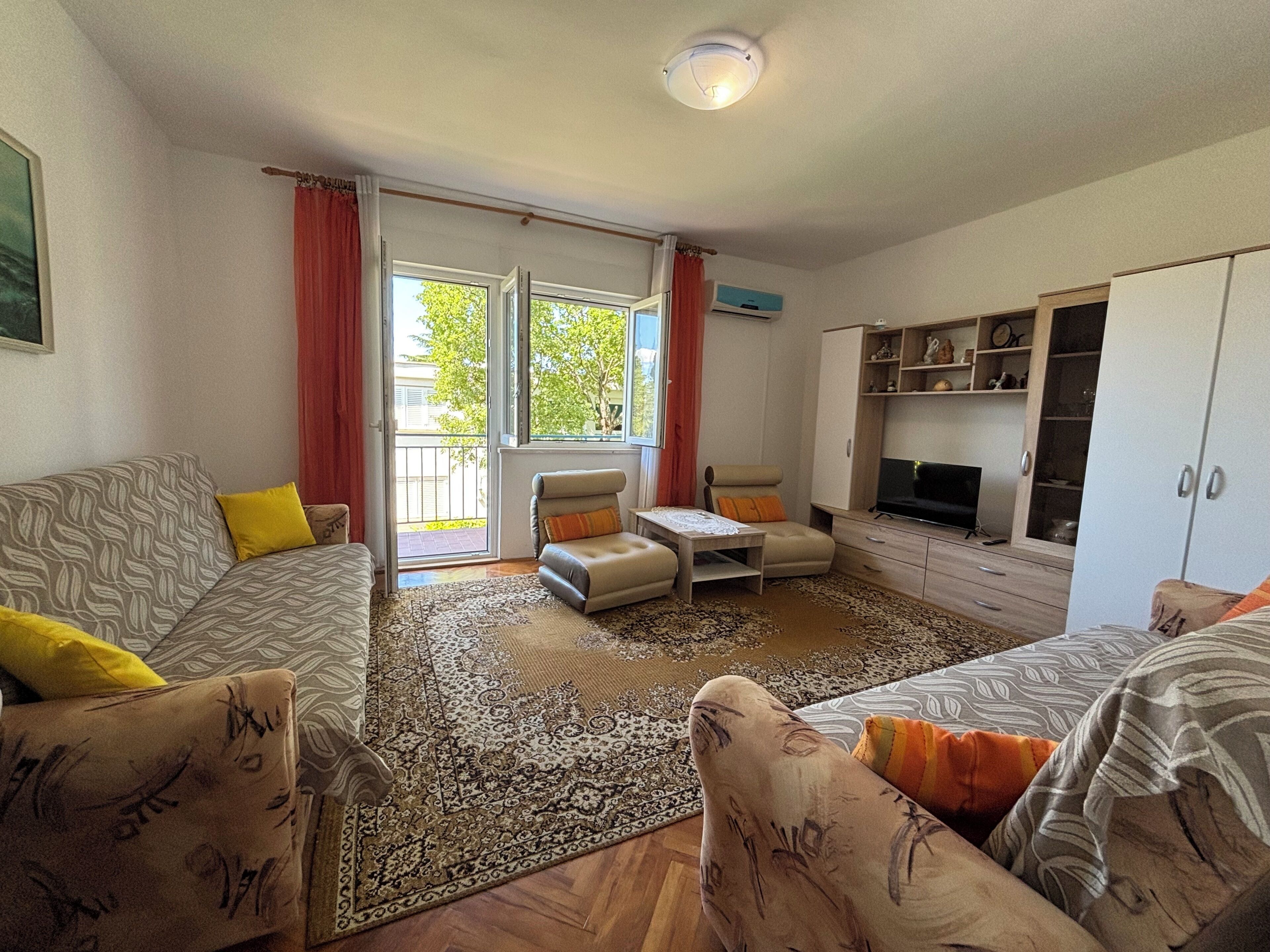 A living room with a sofa, armchairs, a coffee table, and a TV. There is a sliding glass door leading to a balcony.