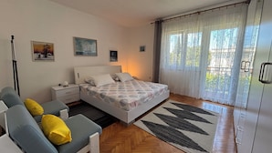 A bedroom with a bed, a sofa, a nightstand, a lamp, a window with curtains, and a herringbone-patterned floor. - Apartman Karlo  (Crikvenica)