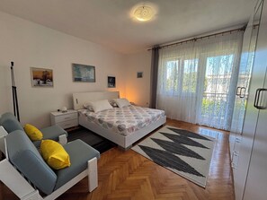 Ein Schlafzimmer mit Bett, Sofa, Nachttisch, Lampe, Fenster mit Vorhängen und einem Fußboden mit Fischgrätenmuster.
