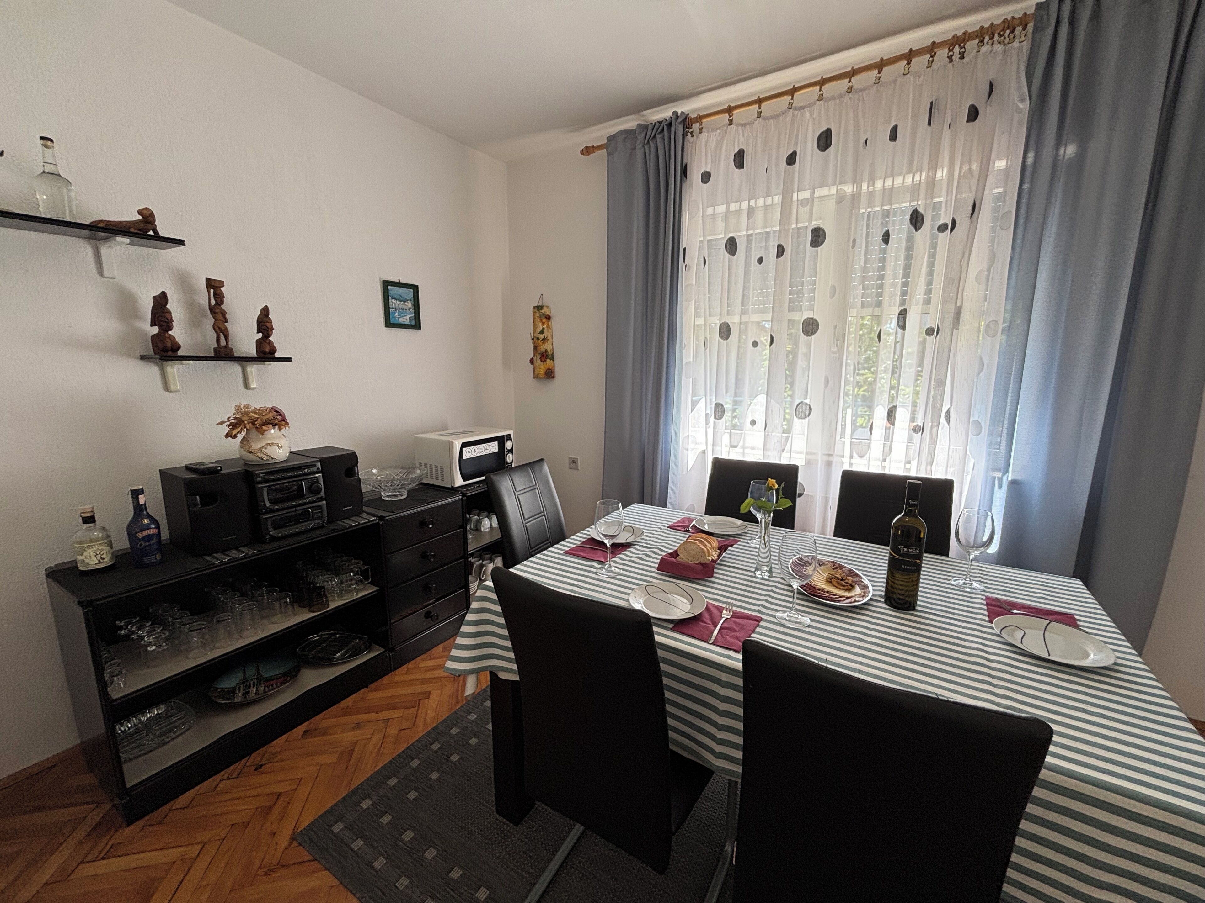 Un coin repas avec une table dressée pour deux personnes, un four à micro-ondes, une petite étagère avec des objets décoratifs et une fenêtre avec des rideaux.