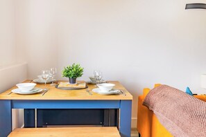 Ein Esstisch ist gedeckt mit Tellern, Gläsern und Besteck, dazu ein kleiner Blumentopf und ein blauer Stuhl mit einer Decke.