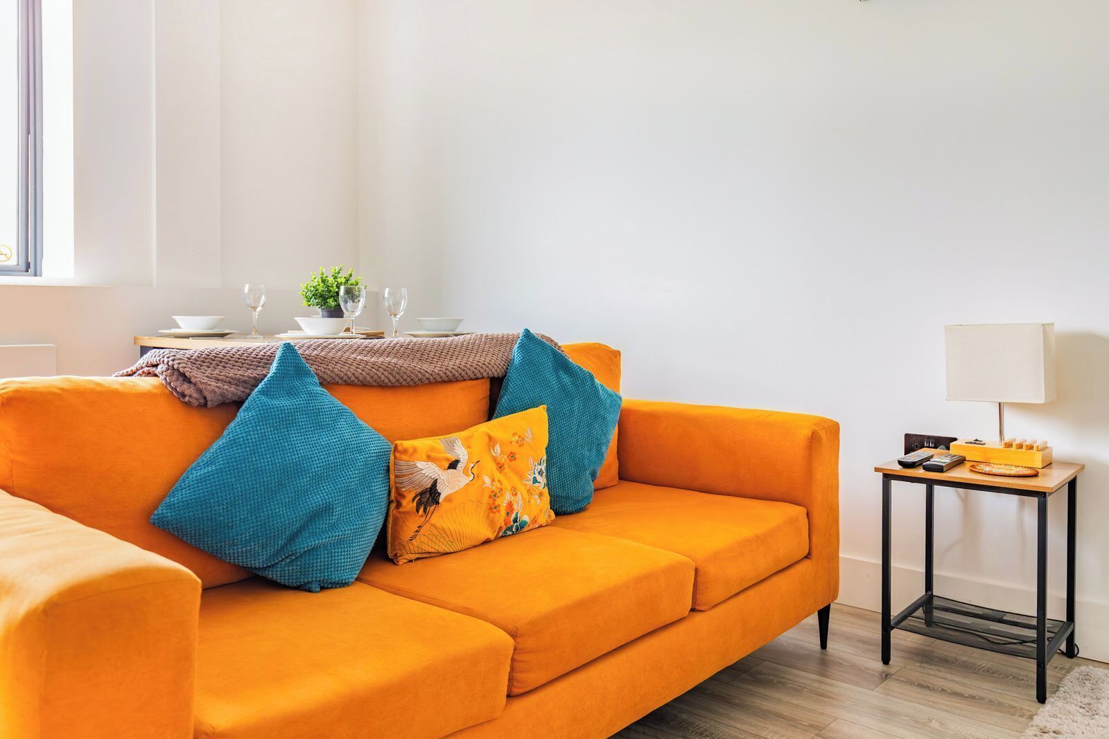 Een feloranje bank met kussens in blauwgroen en geel, een bijzettafeltje met een lamp en een raam met zonwering.