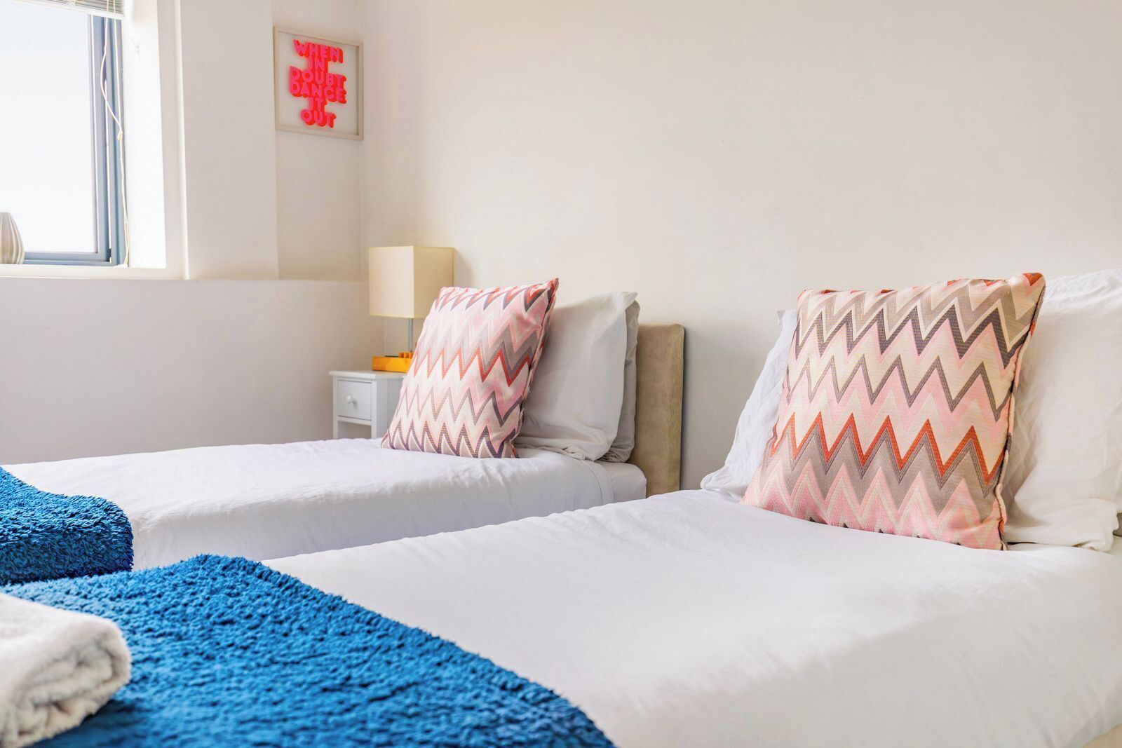 A hotel room with two beds, a nightstand, a lamp, and a window with curtains.