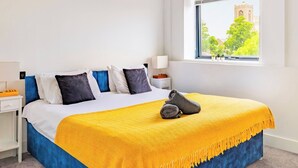 A hotel room with a bed, a nightstand, a lamp, a window with a view of a building and trees. - Stylish Central Location Apartment (Norwich)