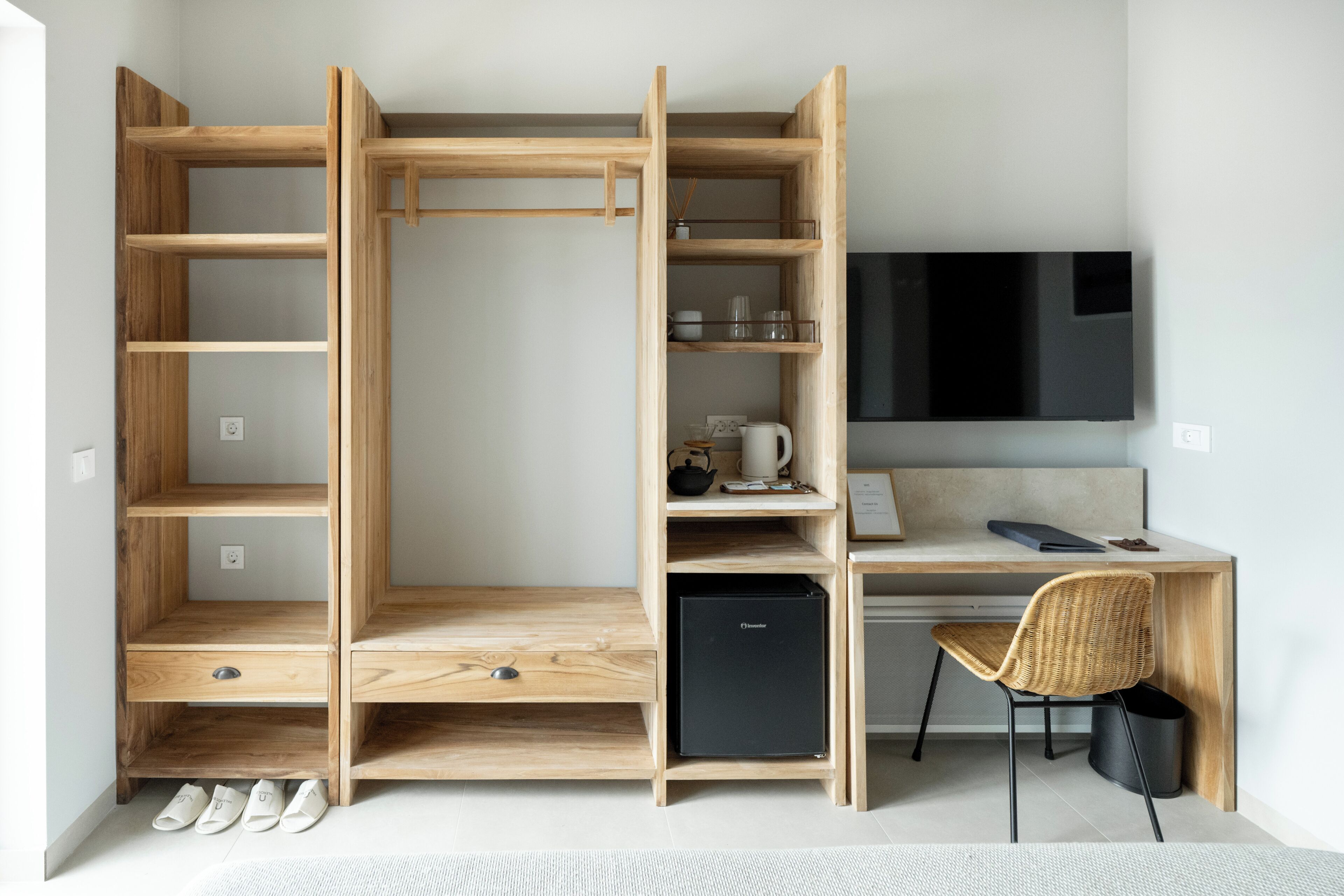 A compact room with a built-in desk, a small black refrigerator, a wicker chair, and a TV mounted on the wall.